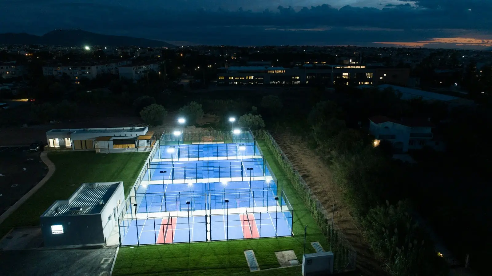 Floodlit padel courts at dusk seen from the air, with the lights falling on the courts